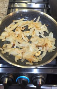 Sliced yellow candy onions and purple garlic caramelizing in a silver skillet on a gas range for Scofield Fruits’ Poblano cream sauce.